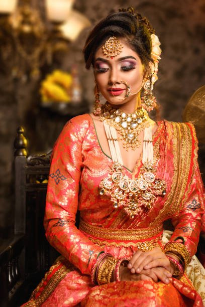 Magnificent young Indian bride in luxurious bridal costume with makeup and heavy jewellery is sitting in a chair in with classic vintage interior in studio lighting. Wedding fashion.