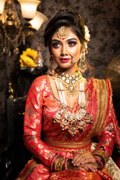 Stunning Indian bride in luxurious bridal costume with makeup and heavy jewellery is sitting in a chair in with classic vintage interior in studio lighting. Wedding lifestyle and fashion.