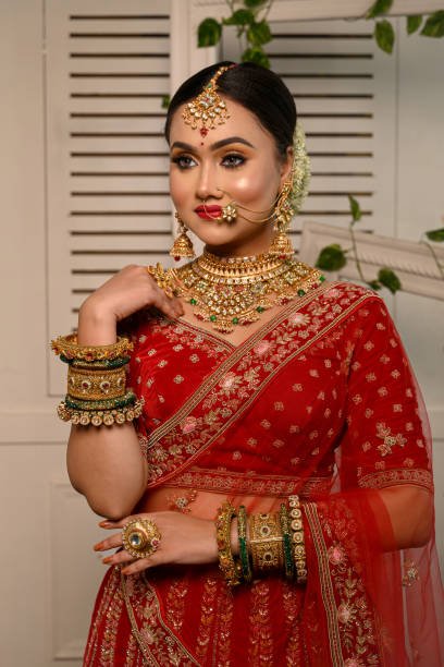 Stunning Indian bride adorned in a traditional red bridal lehenga gracefully wears heavy gold jewelry and a veil and smiles tenderly in studio lighting. Wedding fashion and lifestyle.